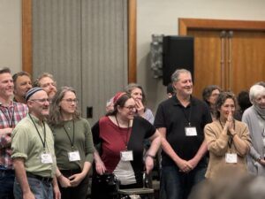 A group of adults stand together indoors, smiling and looking toward something off camera.