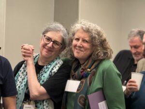 Two older women smiling and holding hands at an indoor event, with people in the background.