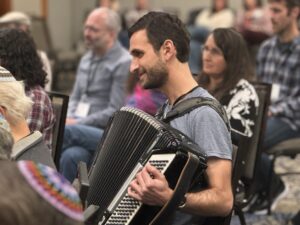 A man plays an accordion in a crowded room, with people seated and listening attentively.