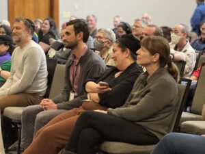A group of people sit closely together in rows, listening and smiling at an indoor event or presentation.
