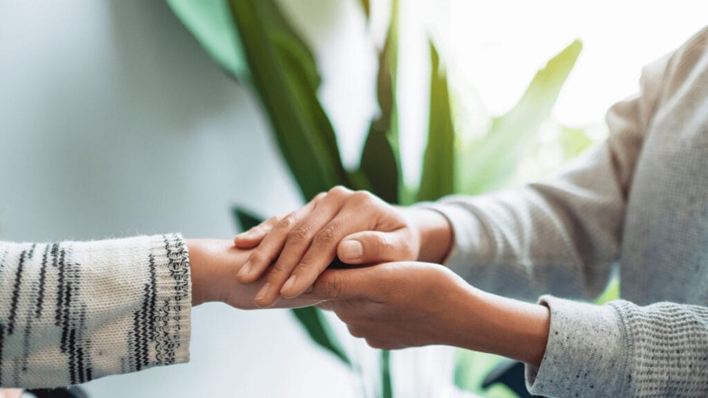 Two people holding hands gently, offering comfort and support, with green plants in the background.