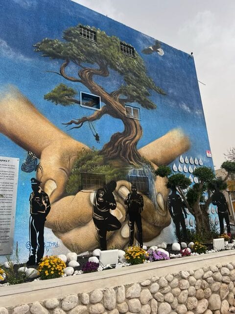A mural of large hands holding a tree, with silhouetted police officers and flowers in front of a blue wall.