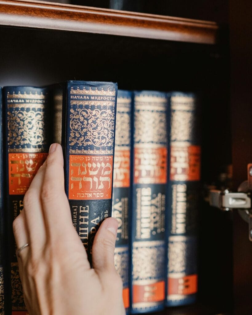 A hand pulling a blue and gold Hebrew book from a shelf with similar books.