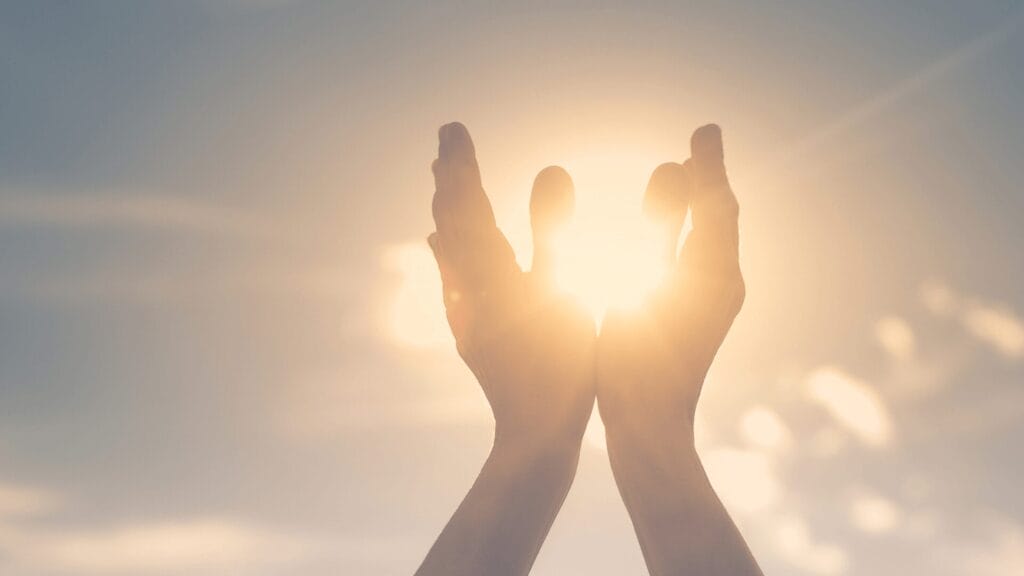 Two hands held up toward the bright sun, with sunlight shining through the space between them.