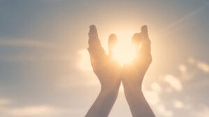 Two hands held up toward the bright sun, with sunlight shining through the space between them.
