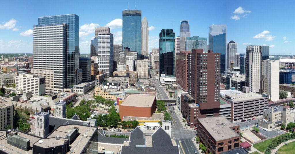 A city skyline with tall modern buildings under a clear blue sky and scattered clouds.