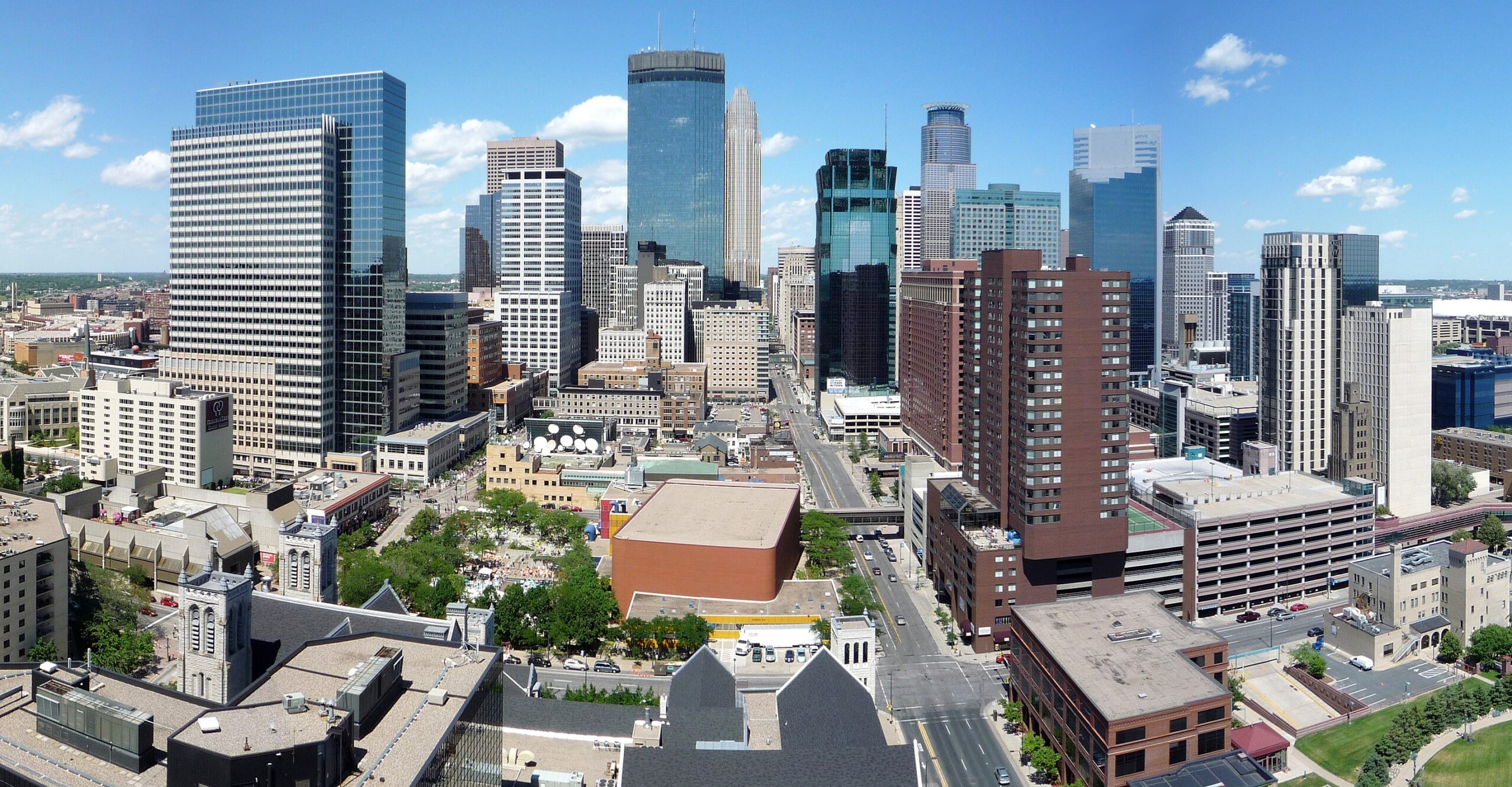 A city skyline with tall modern buildings under a clear blue sky and scattered clouds.