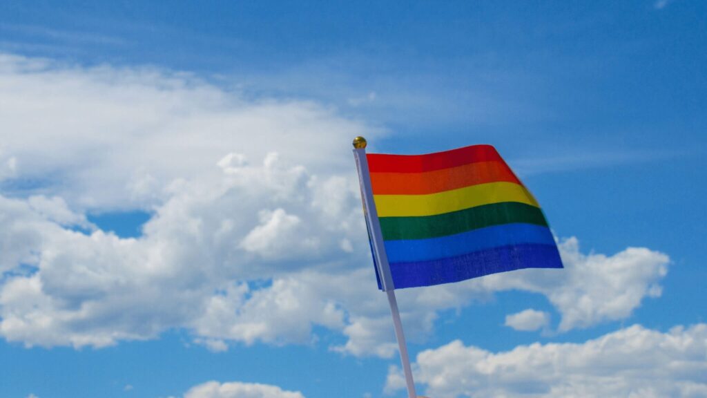 A Pride flag flying against a blue sky with clouds