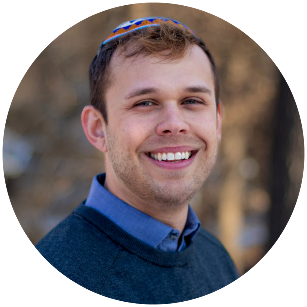 A smiling man in a blue shirt and sweater, wearing a colorful kippah, outdoors with a blurred background.