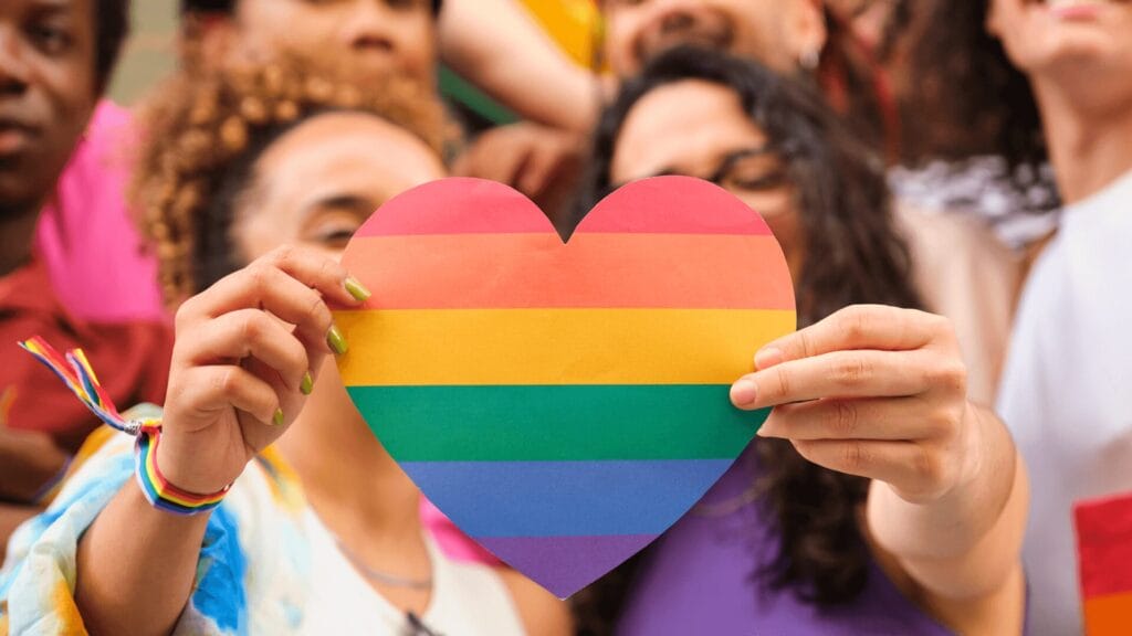 Two people in a crowd holding up a paper heart decorated with rainbow stripes.