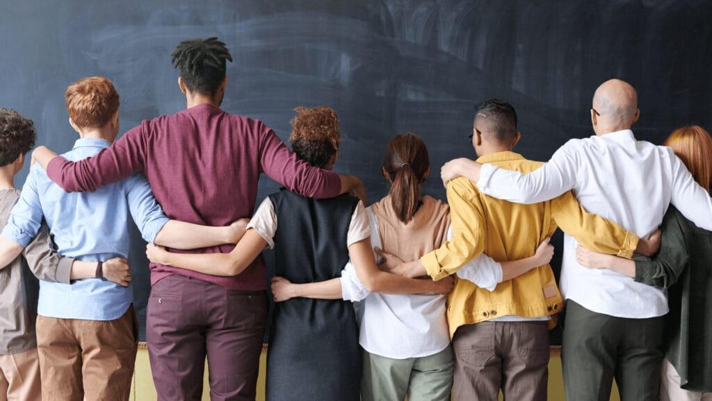 A diverse group of people stand with arms around each other, facing a chalkboard.