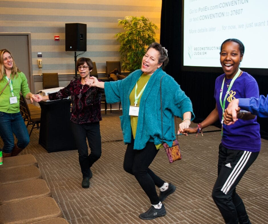 Four women holding hands and dancing in a conference room, smiling and enjoying themselves.
