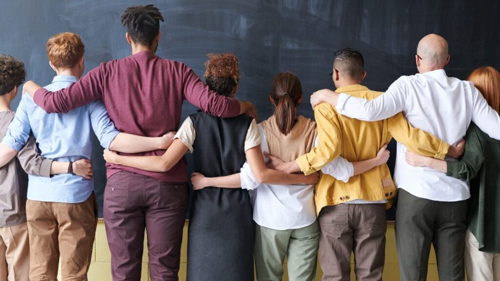A diverse group of people standing in a row with arms around each other, facing a chalkboard.