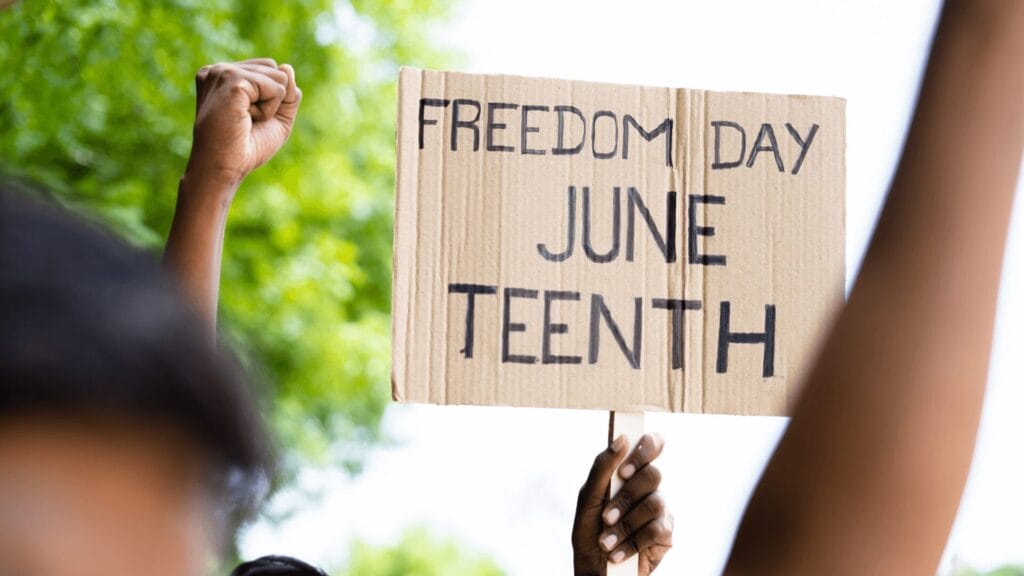 A hand holds a Freedom Day Juneteenth sign; raised fists are visible in the foreground.