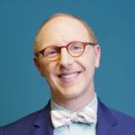 Smiling person with glasses and a striped bow tie, wearing a blue suit, against a teal background.