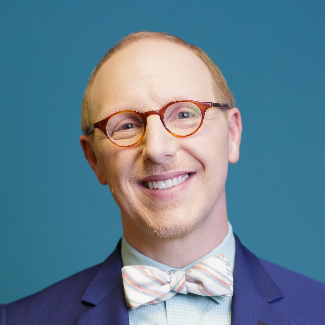 Smiling person with glasses and a striped bow tie, wearing a blue suit, against a teal background.