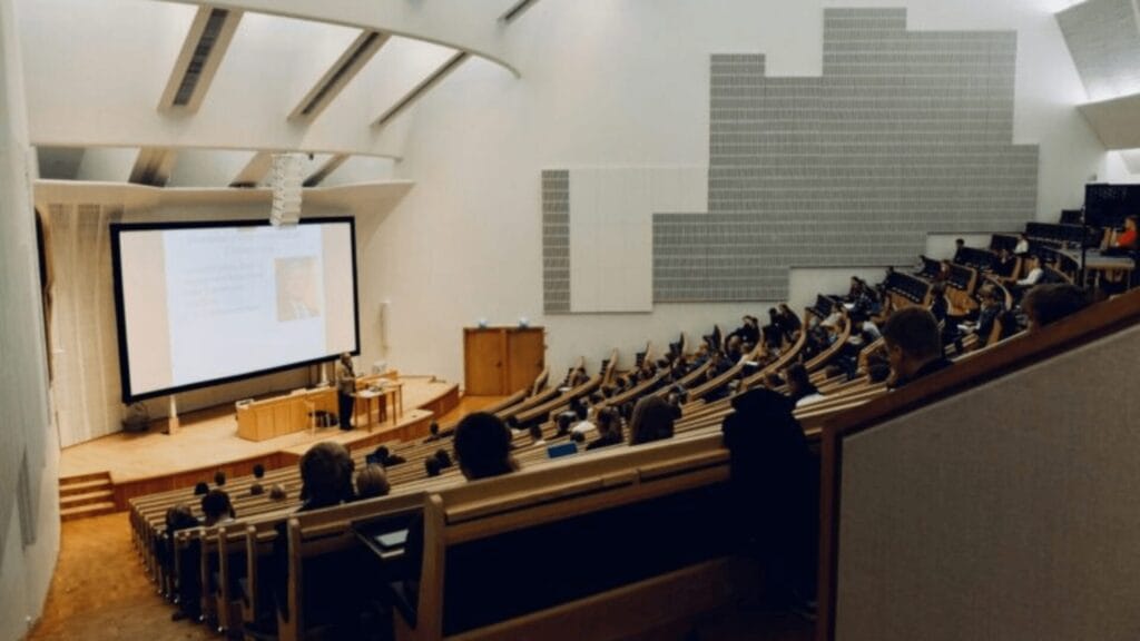Students sit in a large, modern lecture hall watching a presentation on a projector screen.