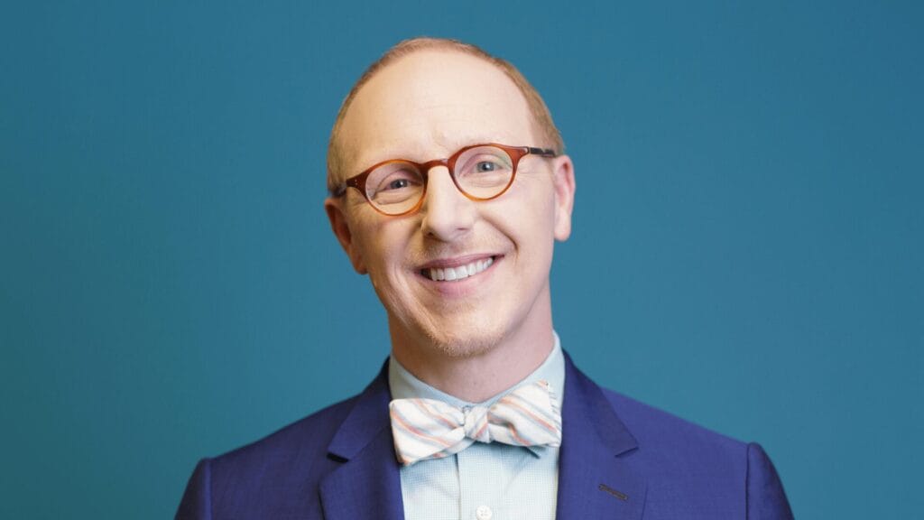 Smiling mam in glasses, a blue suit, and a pastel bow tie, standing against a blue background.
