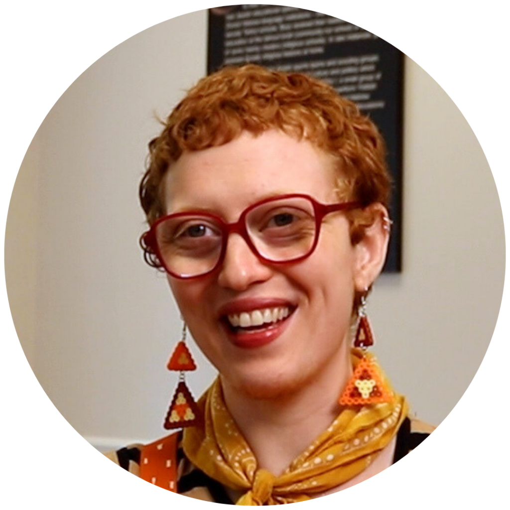 Person with short curly red hair, red glasses, geometric earrings, and a yellow scarf smiling.