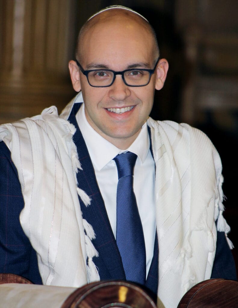 A smiling man with glasses wearing a suit, tie, and a white prayer shawl.