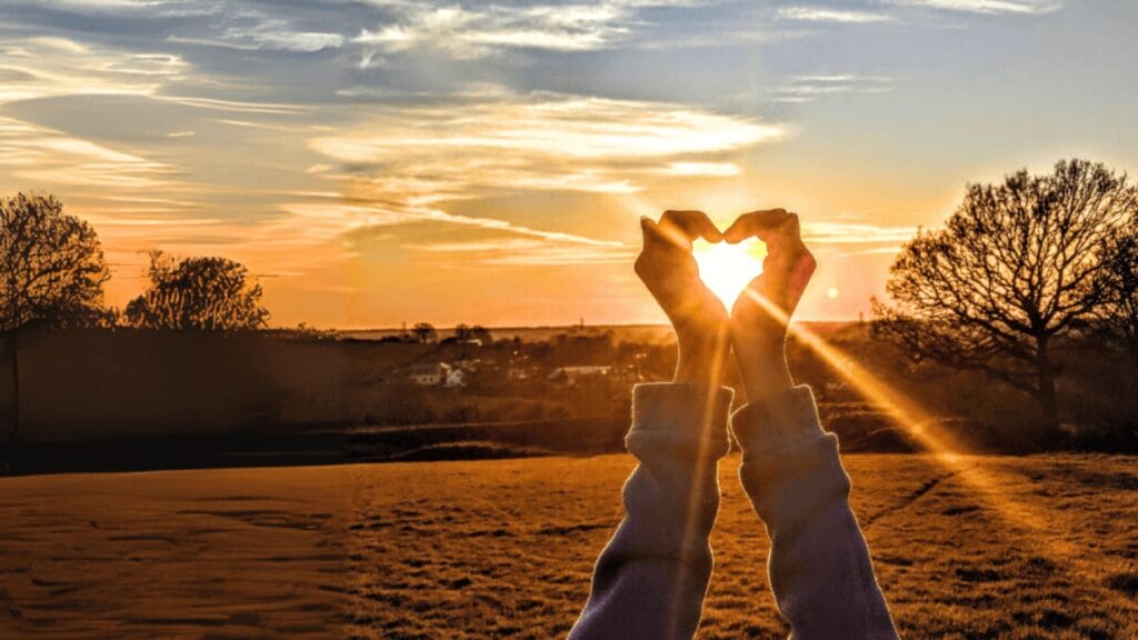 Hands form a heart shape around the sun at sunset in a field, with trees and clouds in the background.