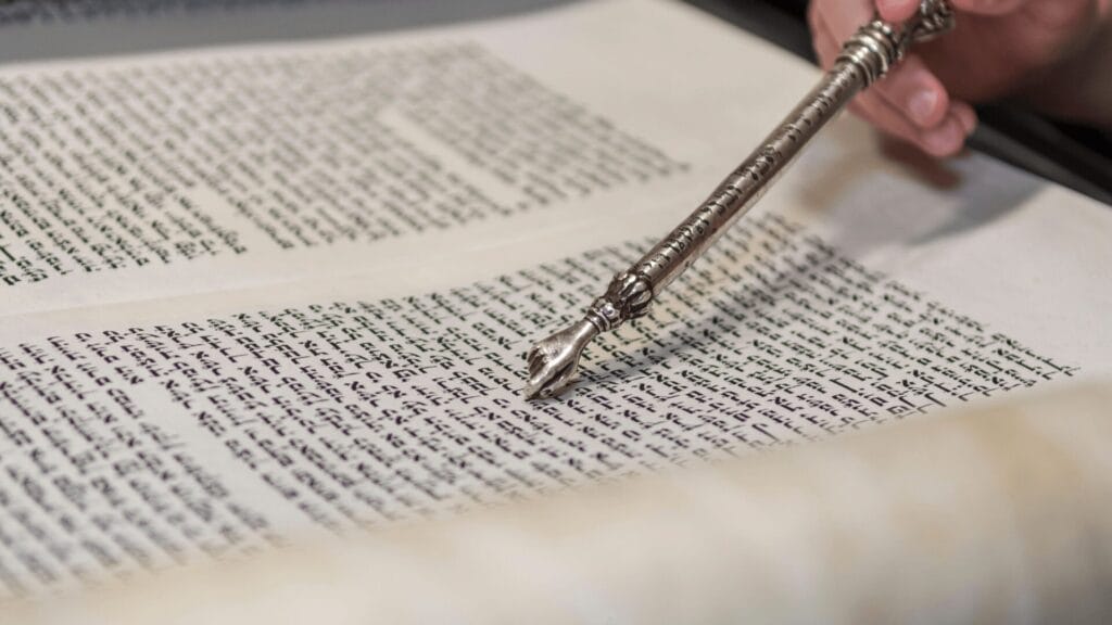 A hand uses a silver pointer to follow Hebrew text on an open Torah scroll.