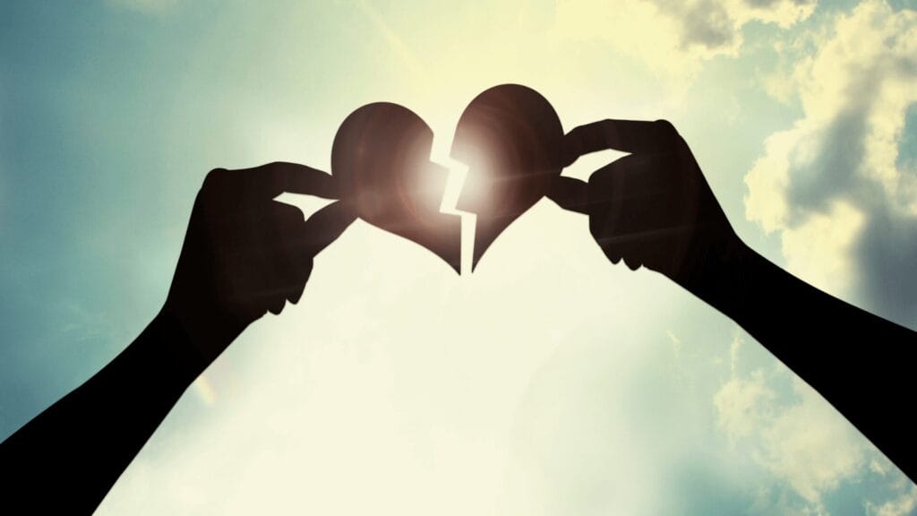 Two hands hold pieces of a broken heart together, creating a heart shape against a bright sky.
