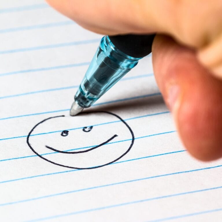 A hand draws a smiling face with a pen on lined notebook paper.