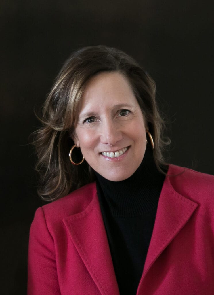 Smiling woman with wavy brown hair in a red blazer and black turtleneck against a dark background.