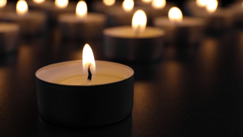 A group of small tea light candles burning in the dark, with one candle in sharp focus in the foreground.