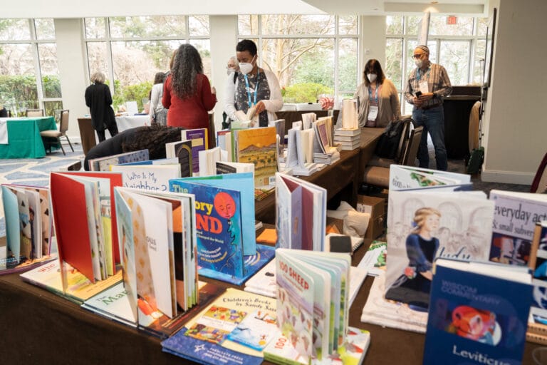 Books displayed on a table at an indoor event, with people browsing and talking in the background.