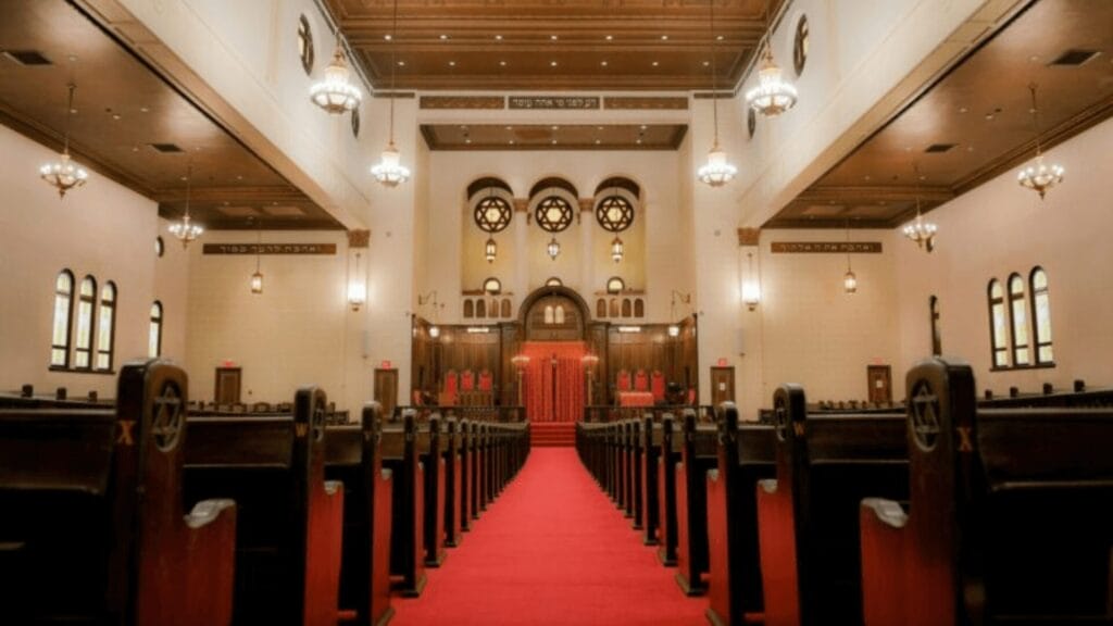 Interior of a synagogue with wooden pews, red carpet aisle, and ornate stained glass windows at the front.