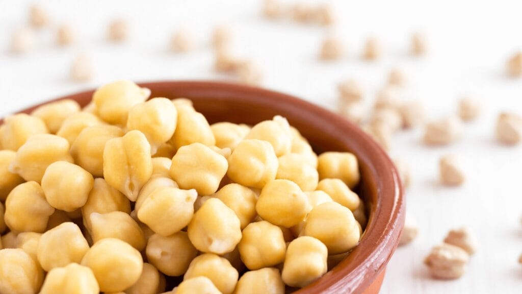 A brown bowl filled with chickpeas, with more chickpeas scattered in the background.