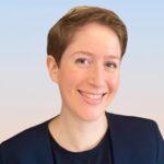 Person with short brown hair, smiling, wearing a navy blazer and black shirt against a light gradient background.