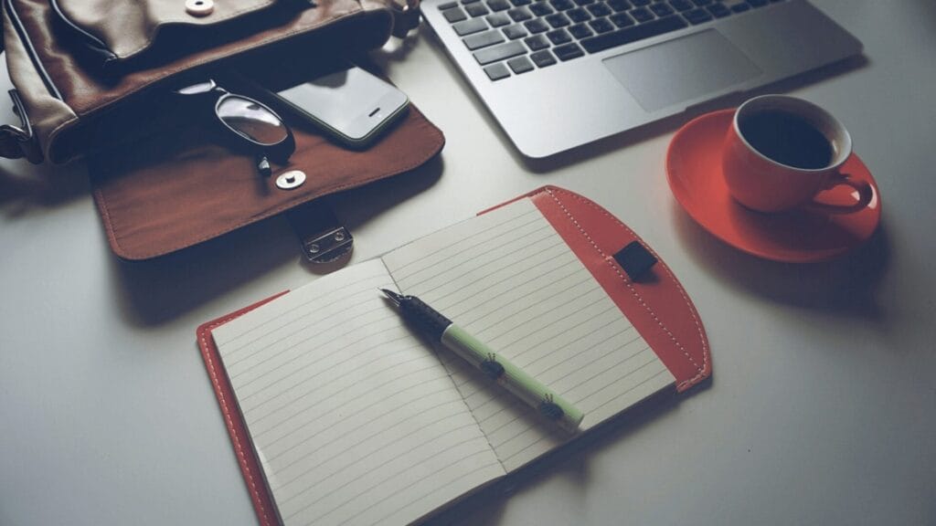Open notebook with pen, laptop, coffee cup, glasses, and bag on a desk.