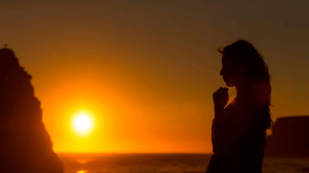 A silhouette of a person standing by the ocean at sunset, hands clasped, with cliffs in the background.