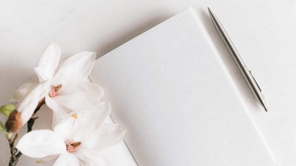 Open blank notebook with a silver pen and white flowers on a white surface.
