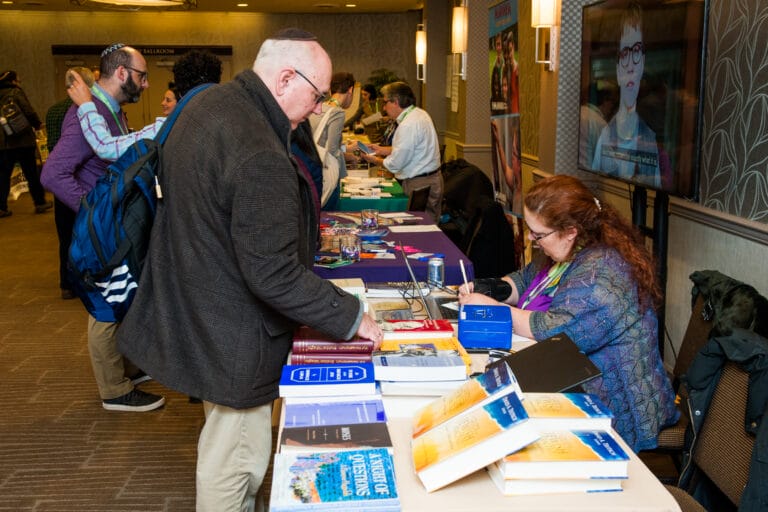 People browsing books and speaking to vendors at a conference or book fair table inside a hotel.