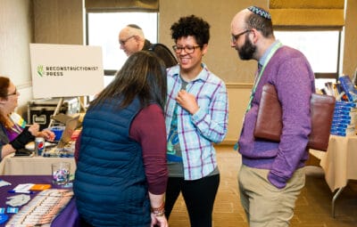 Three people smile and chat near a table at a conference with a Reconstructionist Press sign.