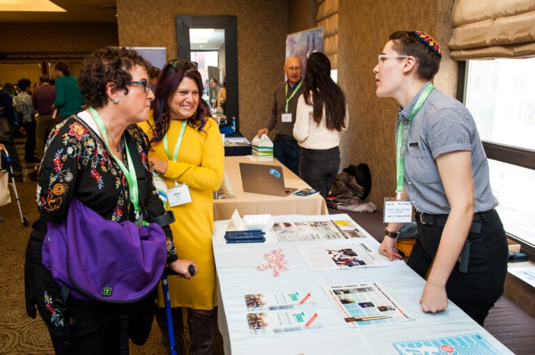 Three women converse at an information booth during a conference, pamphlets and candies on the table.