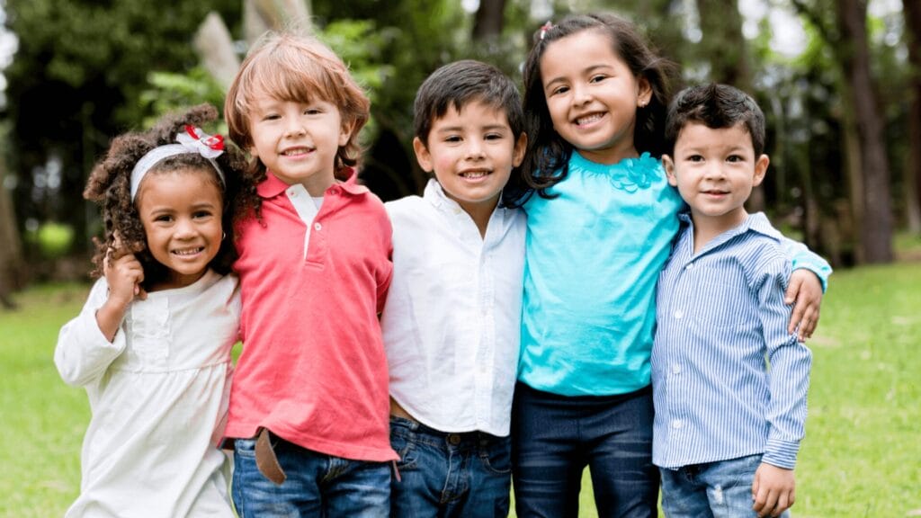 Five smiling children stand arm in arm outdoors, posing together in a grassy park.