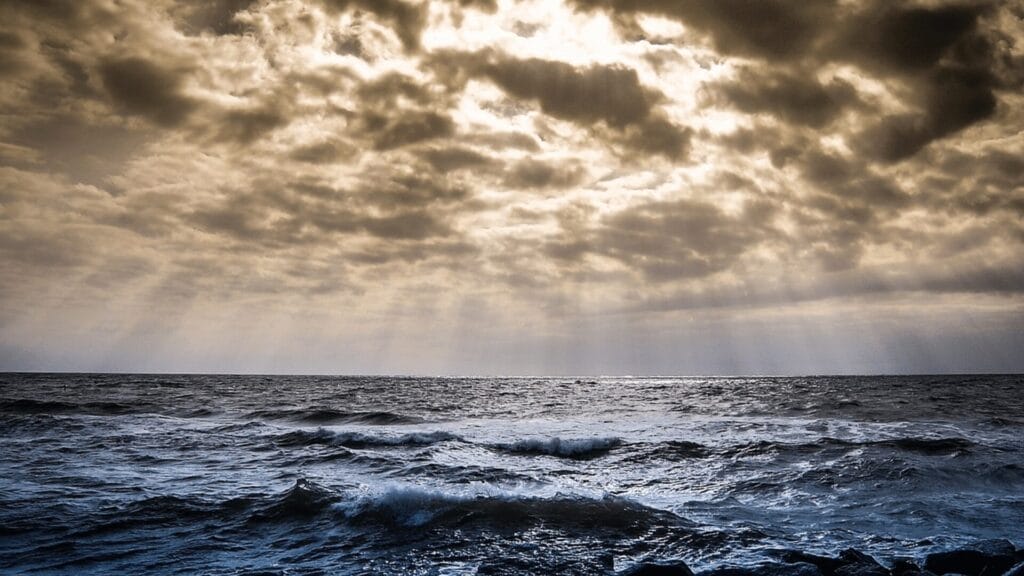 Waves crash under a cloudy sky with sun rays streaming through, creating a dramatic seascape.
