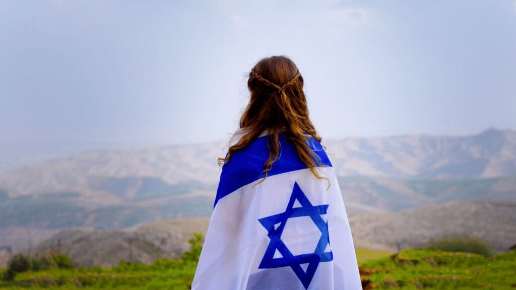 A person draped in an Israeli flag stands outdoors, facing distant mountains under a cloudy sky.