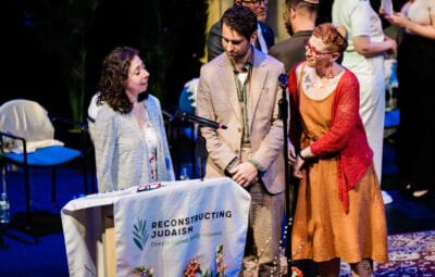 Three people speak at a podium with a Reconstructing Judaism banner during a formal event.
