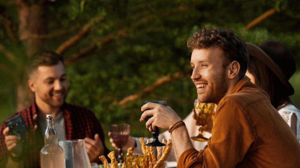 Three friends sit outdoors at a table, smiling and holding drinks, with greenery in the background.