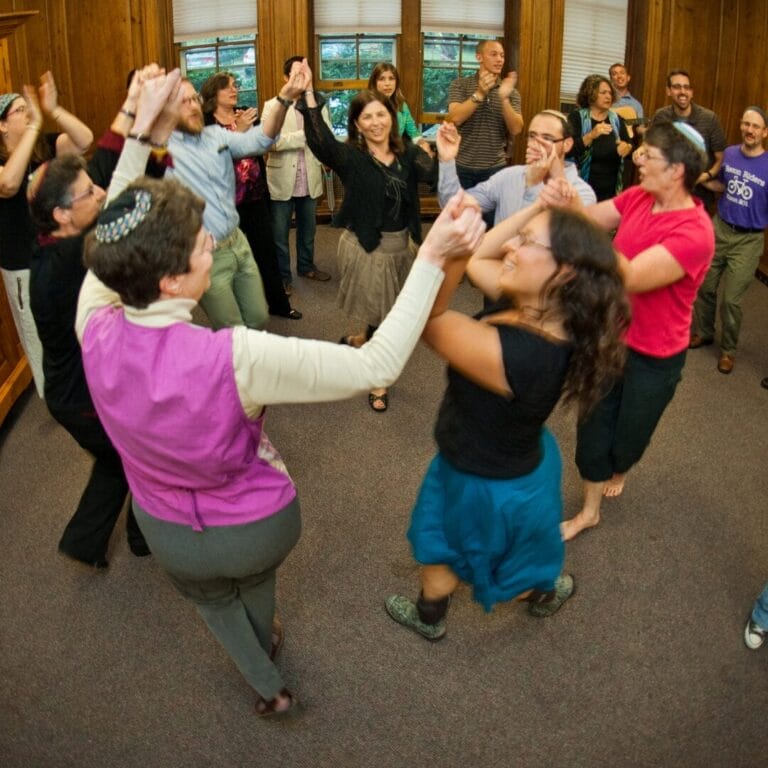 A group of people dance in a circle indoors, smiling and holding hands in a lively, joyful atmosphere.