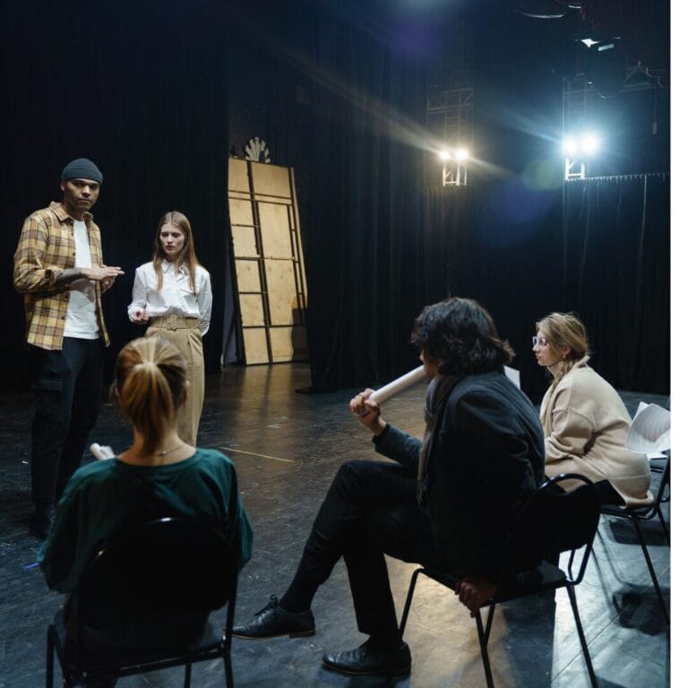 Two people stand onstage talking while three others sit and watch in a dimly lit theater setting.