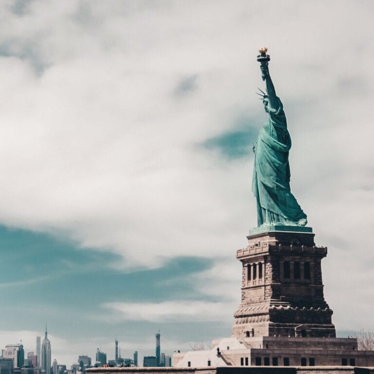The Statue of Liberty stands tall against a cloudy sky with the New York City skyline in the background.