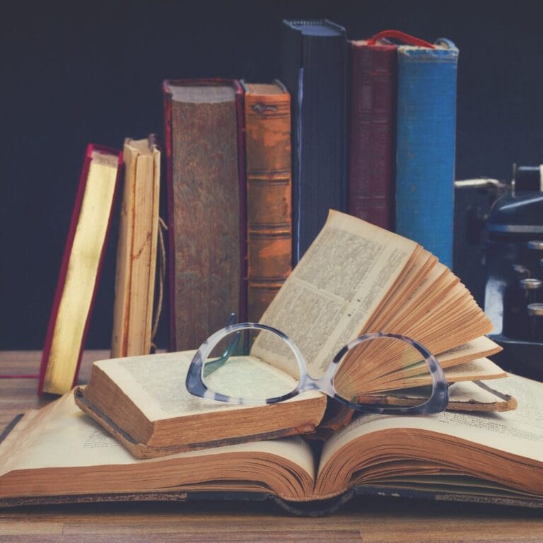A pair of glasses rests on an open book, surrounded by stacks of old books on a wooden table.