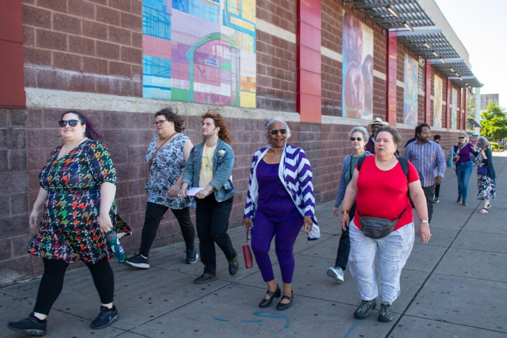 A group of people walk together on a city sidewalk beside a brick building with colorful murals.